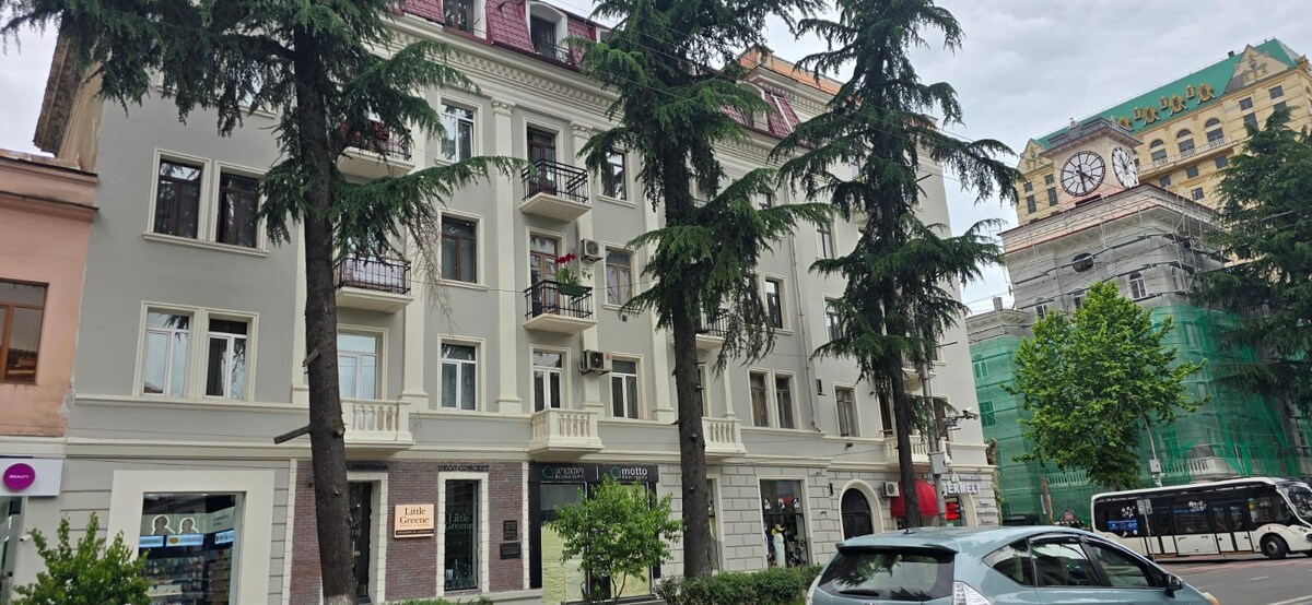 A modern multifamily building is presented, featuring balconies and large windows framed by tall trees. The light-colored façade contrasts with the greenery around. A bus is visible along the street, and a historic building is seen in the background, partially covered by scaffolding.