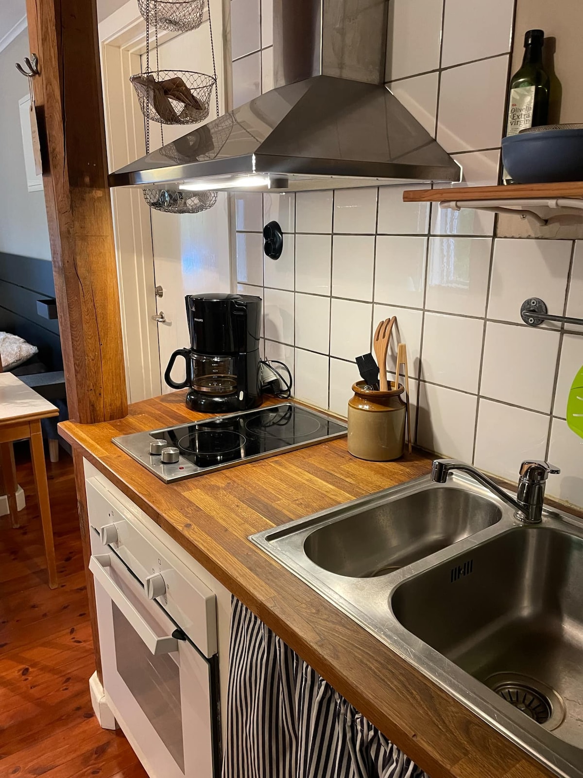 A functional kitchen space is highlighted with wooden countertops and stainless steel appliances. A double sink is positioned next to a two-burner stovetop, while a coffee maker and utensils are visible on the countertop. A warm, inviting atmosphere is noted with natural wood accents and white tiles.