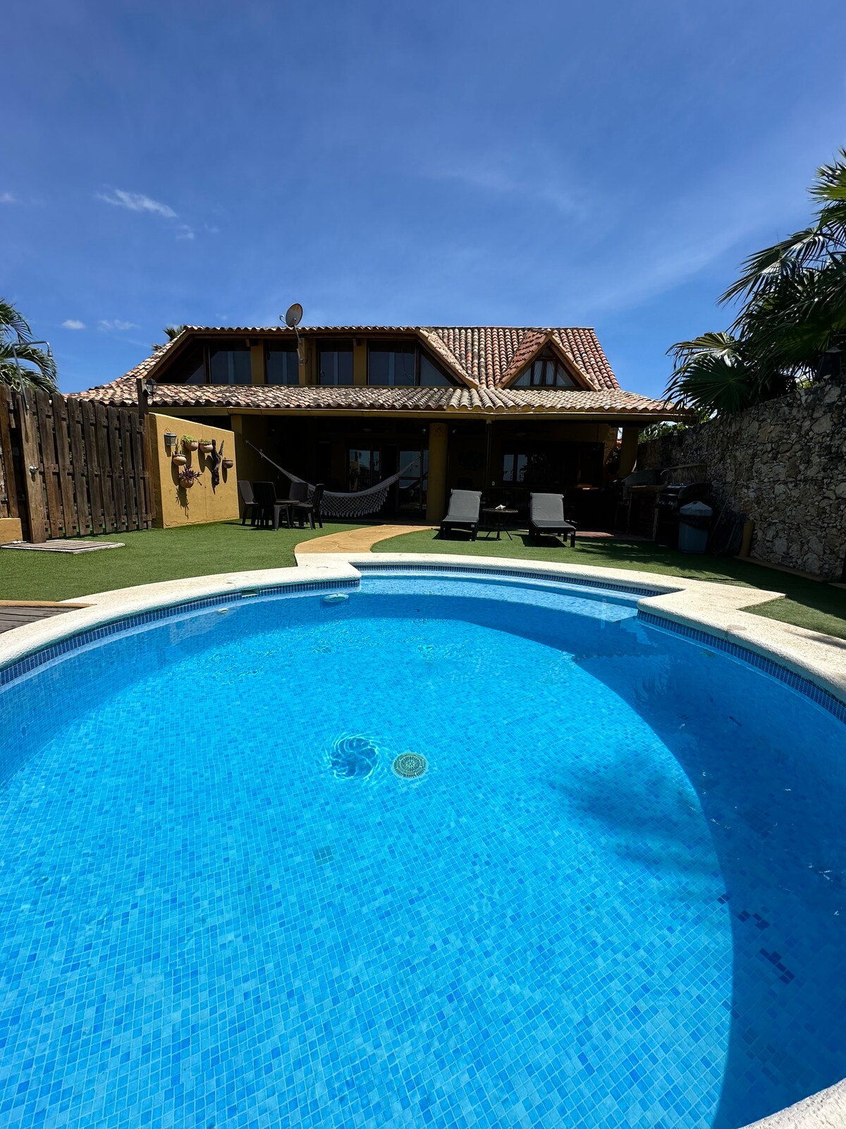 A sparkling blue pool is centered in the foreground, surrounded by a spacious deck. The charming two-story house with a tiled roof is visible in the background, complemented by palm trees and a shaded patio area with lounge chairs.