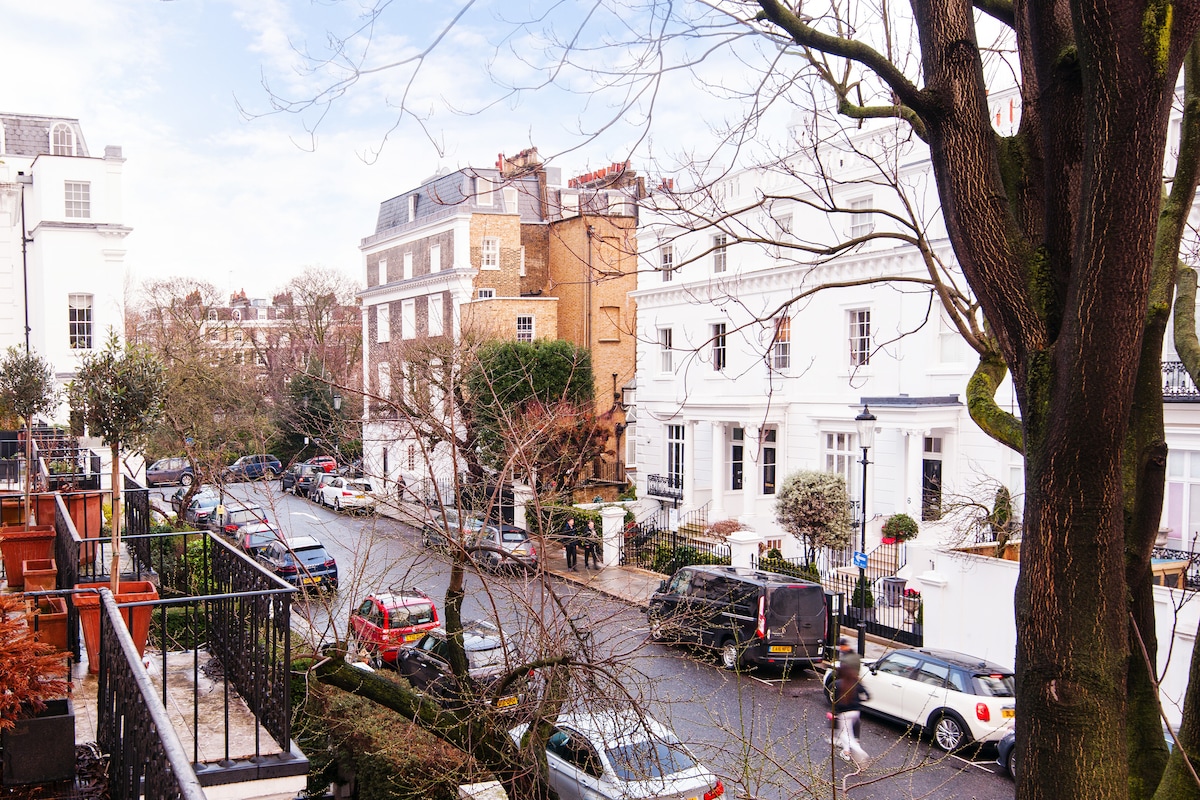 A view of a tranquil street showcasing a mix of elegant white and brick buildings. Lush trees line the road, creating a natural canopy. Several parked cars are visible along the street, offering a sense of a quiet residential area.