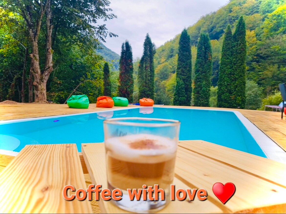A clear swimming pool is surrounded by wooden decking and lush green trees. Colorful bean bags in shades of orange and green are positioned nearby, providing a cozy seating area. A glass of coffee is placed on the edge of the deck, evoking a tranquil atmosphere.