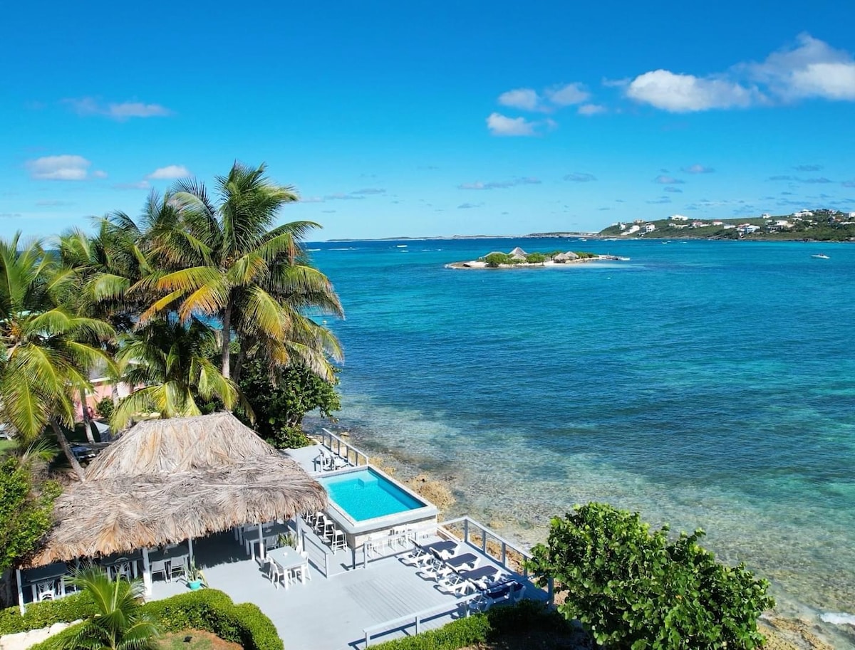An oceanfront view highlights the sparkling turquoise waters and lush greenery surrounding the property. A thatched-roof pavilion with lounge chairs sits adjacent to the pool, providing a relaxed atmosphere. Small islands can be seen in the distance under a clear blue sky.