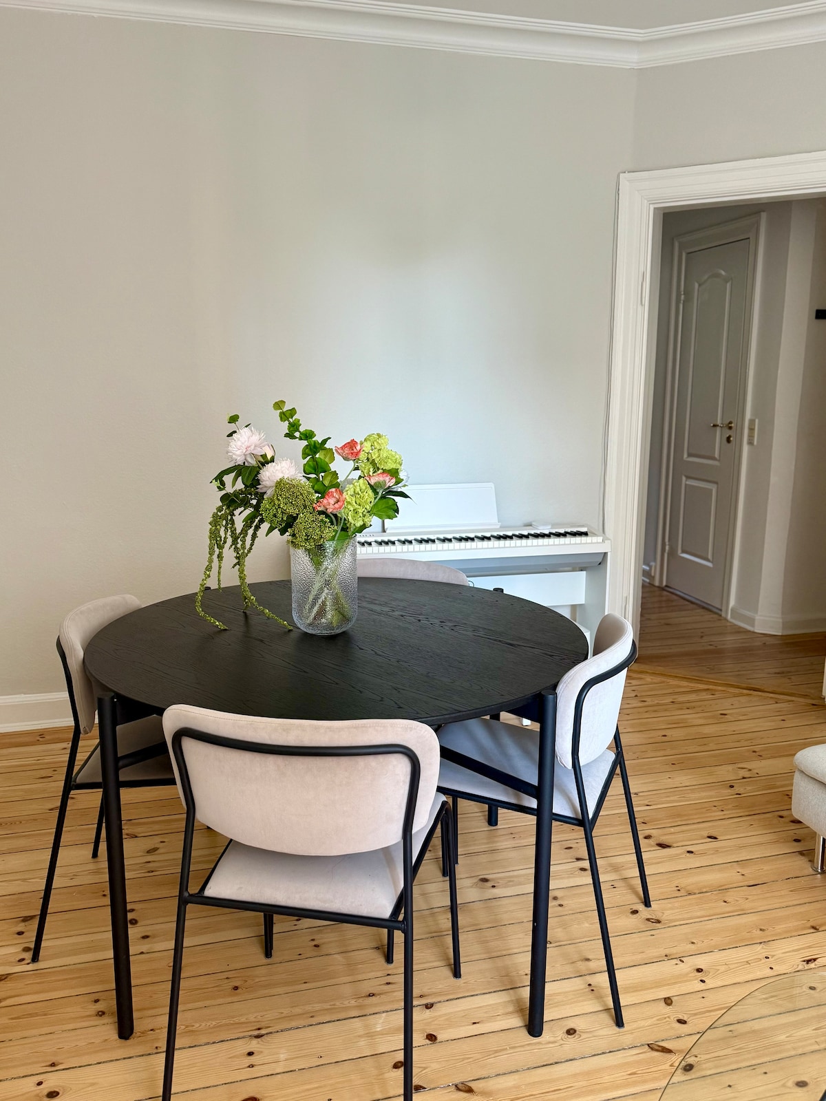 A round dining table with four chairs is positioned in a well-lit area. A vase filled with fresh flowers sits at the center of the table. In the background, a piano is visible, suggesting a creative space. The light wooden floor complements the decor.