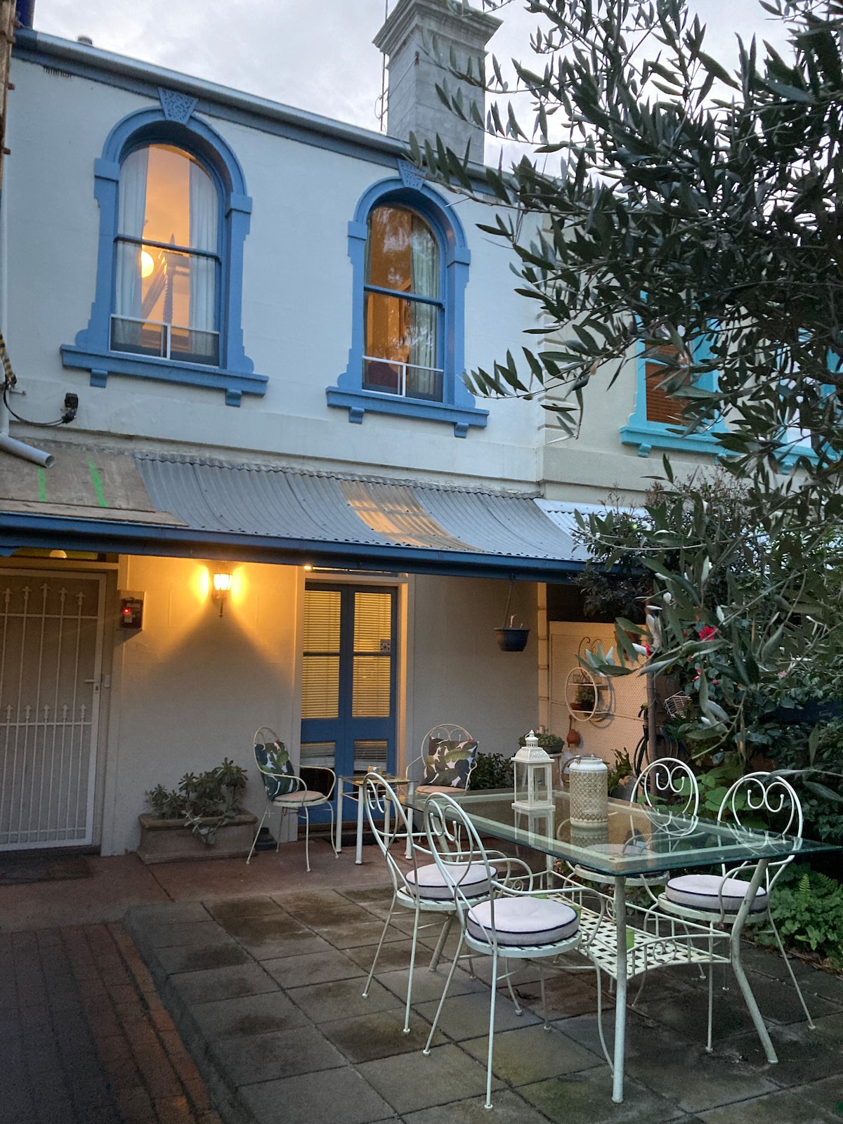 A charming outdoor seating area is showcased, featuring a white metal table surrounded by matching chairs. Potted plants add greenery, while the blue door and windows of the Victorian terrace are visible in the background, enhancing the inviting ambiance of the space.