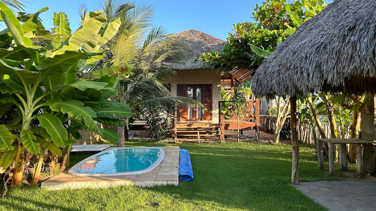 A relaxing outdoor space features a small pool surrounded by lush greenery and tropical plants. A thatched-roof gazebo is positioned nearby, and a wooden deck can be seen in the background, offering comfortable seating options. Bright blue skies complement the inviting environment.
