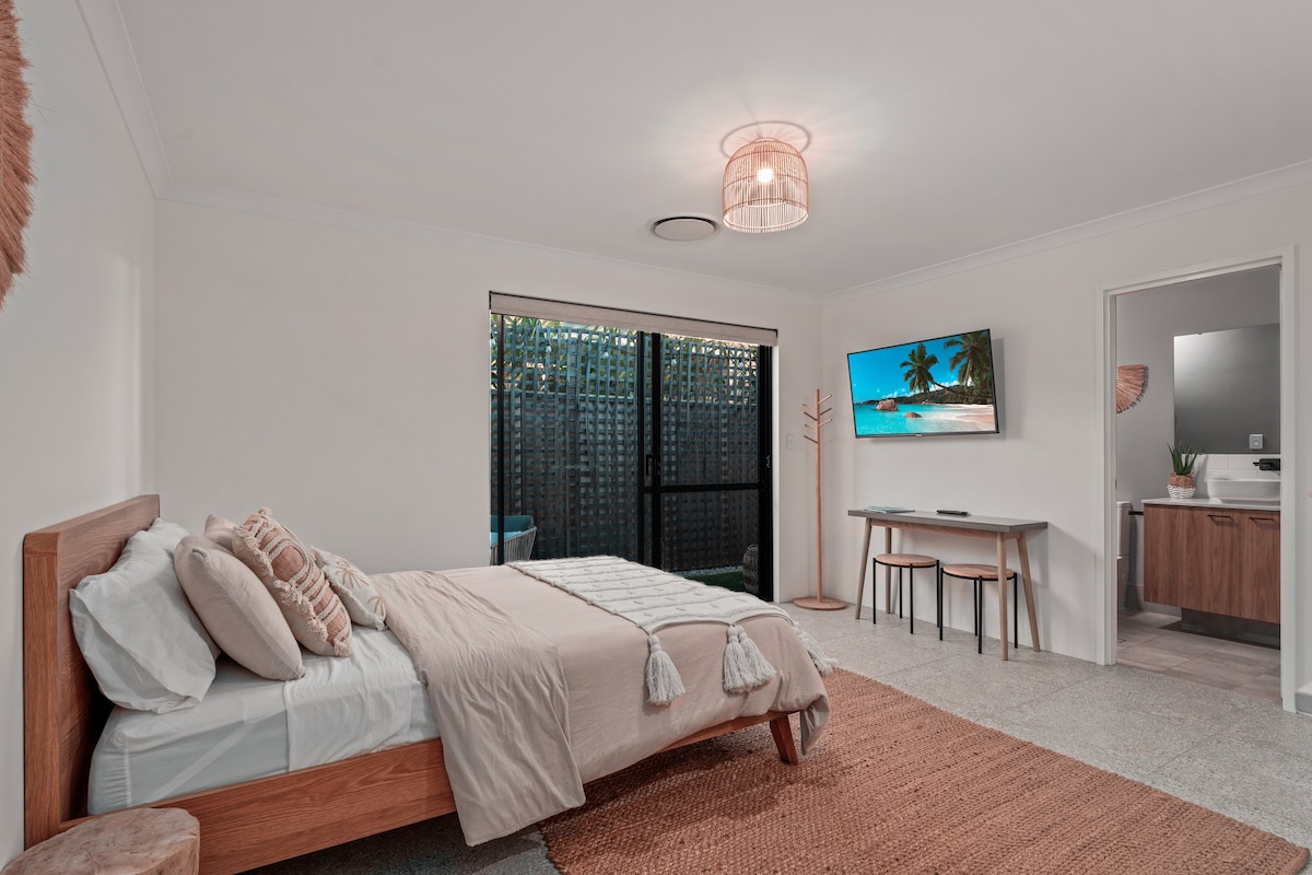 A spacious studio is presented with a queen bed adorned with textured pillows. A wall-mounted TV displays a serene beach scene. Natural light filters through sliding glass doors accessing a private courtyard. A desk and chair are positioned nearby, offering a functional workspace.