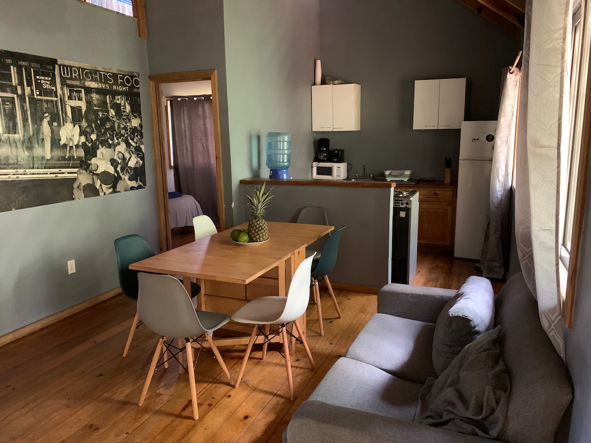A dining area features a wooden table surrounded by six chairs, with a small fruit centerpiece. A comfortable gray sofa is positioned nearby. Kitchen appliances, including a refrigerator and microwave, are visible in the background, alongside a water dispenser and cupboards.