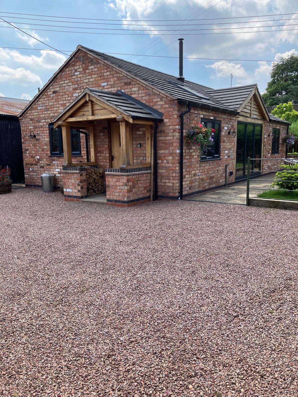 A single-story building constructed with reclaimed bricks features a welcoming facade with a wooden entrance porch. The gravel driveway provides access to the property, surrounded by landscaped areas and large windows reflecting the natural light.