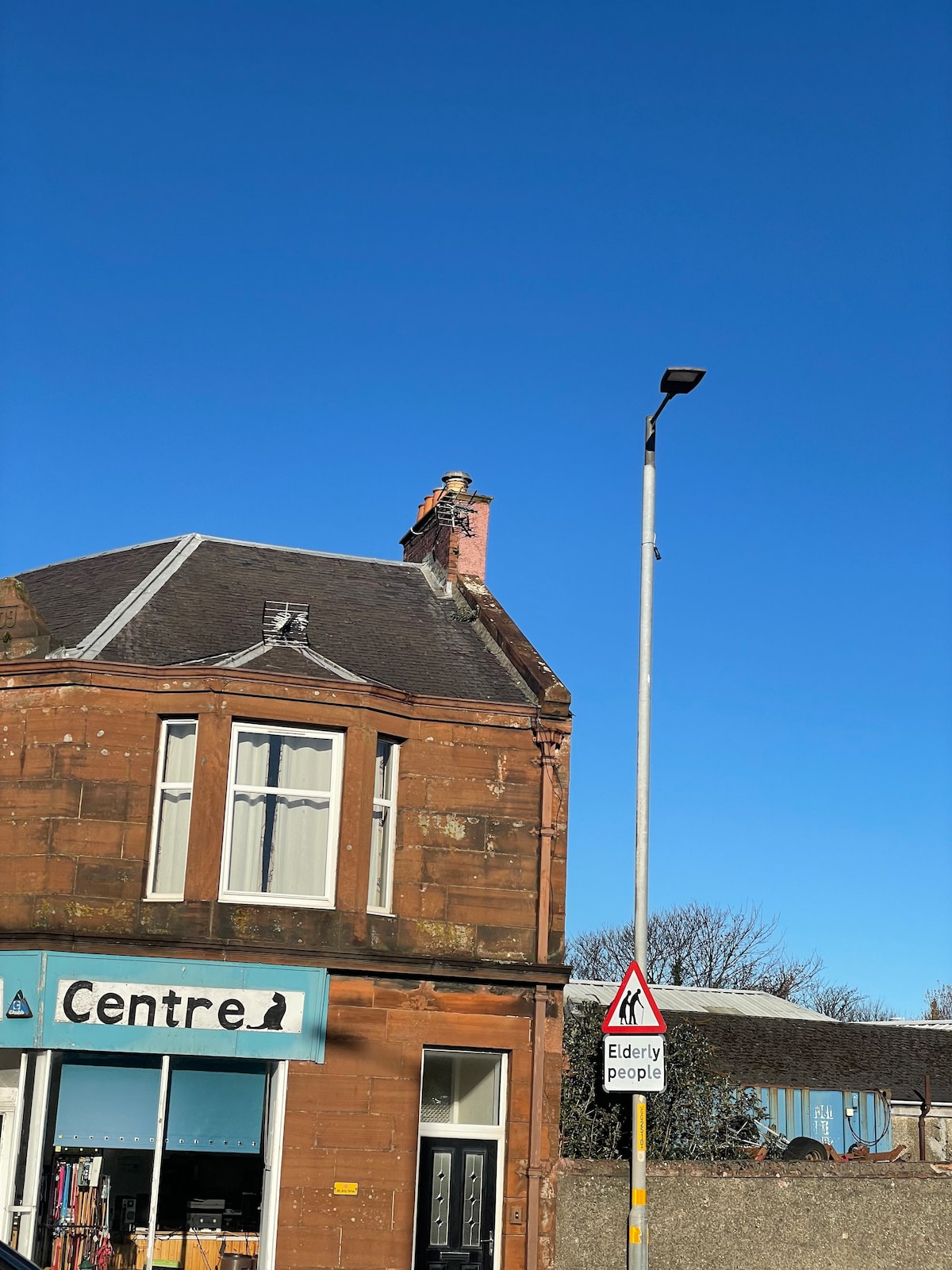 A corner building with a warm orange-brown facade is shown against a clear blue sky. The structure features an angled roof with a chimney. A street sign warning of elderly people is visible nearby, highlighting the community-oriented atmosphere.