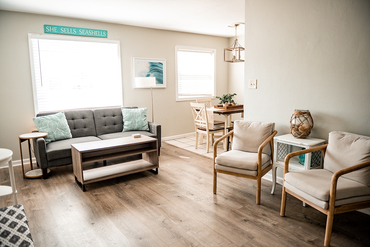 A bright living area is featured, displaying a gray sofa with decorative pillows, a wooden coffee table, and two armchairs. Natural light enters through windows dressed with light curtains. A dining table is positioned in the corner, complemented by a decorative lamp and wall art.