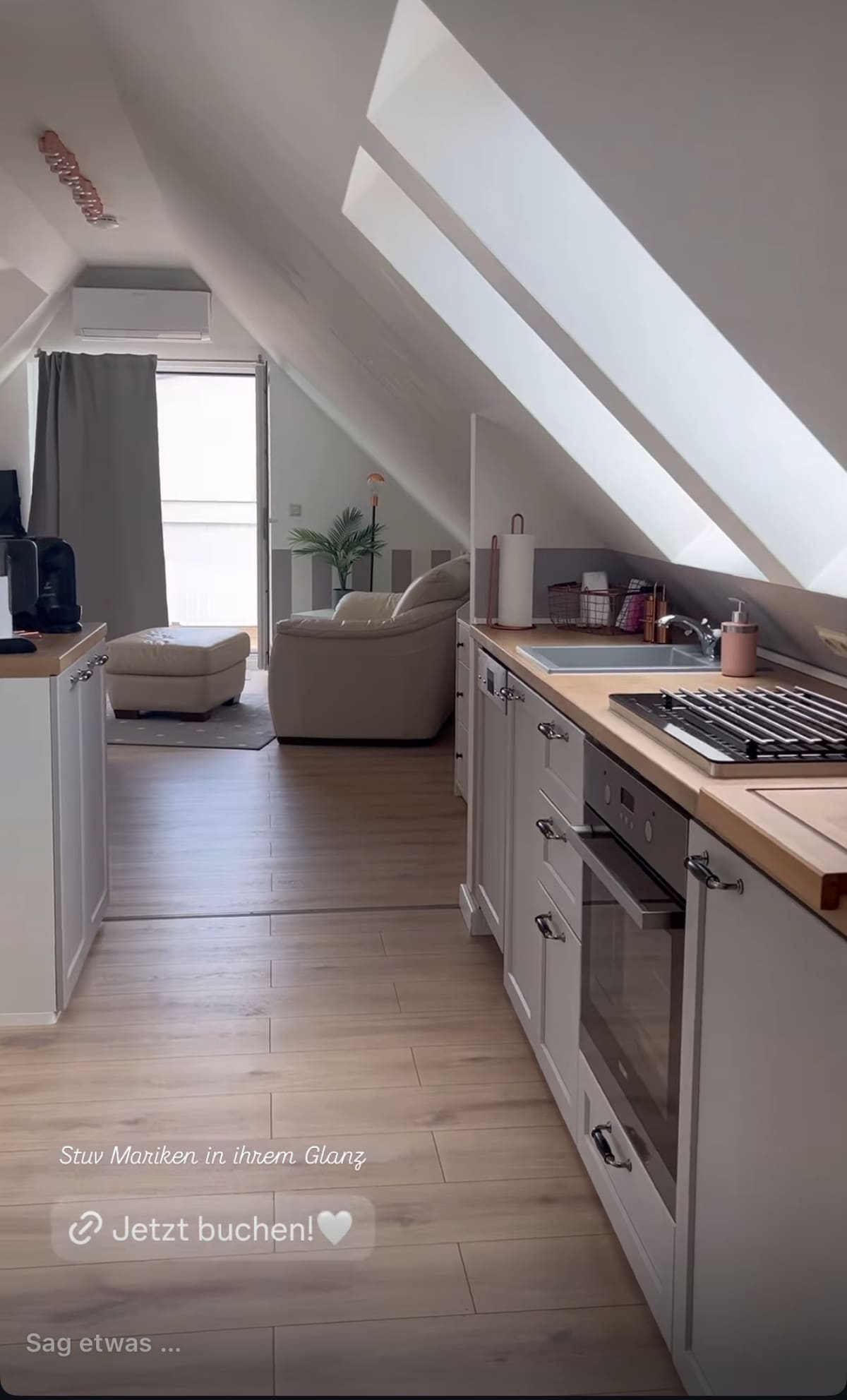 The interior features a clean and modern kitchen equipped with a built-in oven and gas stove. Natural light fills the space from the large window beside a cozy seating area, which includes a comfortable armchair. The white cabinetry and warm wooden accents create a light atmosphere.