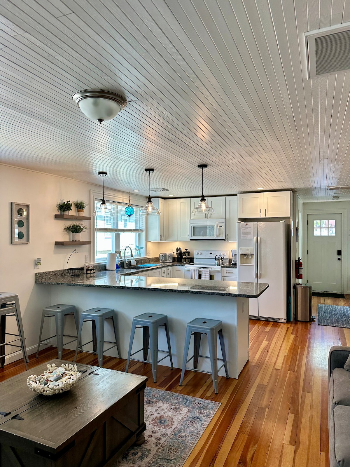 An open-concept kitchen features granite countertops and modern white cabinetry. Bar stools are situated at the kitchen island, providing seating. Stainless steel appliances are visible, and natural light enters through the nearby door. The hardwood floor adds warmth to the space.