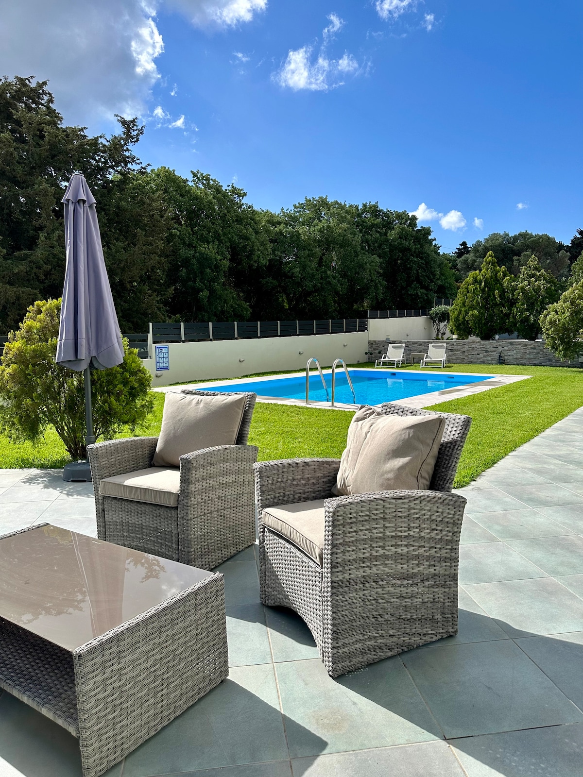 An outdoor seating area is showcased, featuring two comfortable armchairs with neutral cushions beside a small glass-top table. The well-maintained lawn leads to a blue swimming pool, surrounded by greenery and framed by a sunny sky.