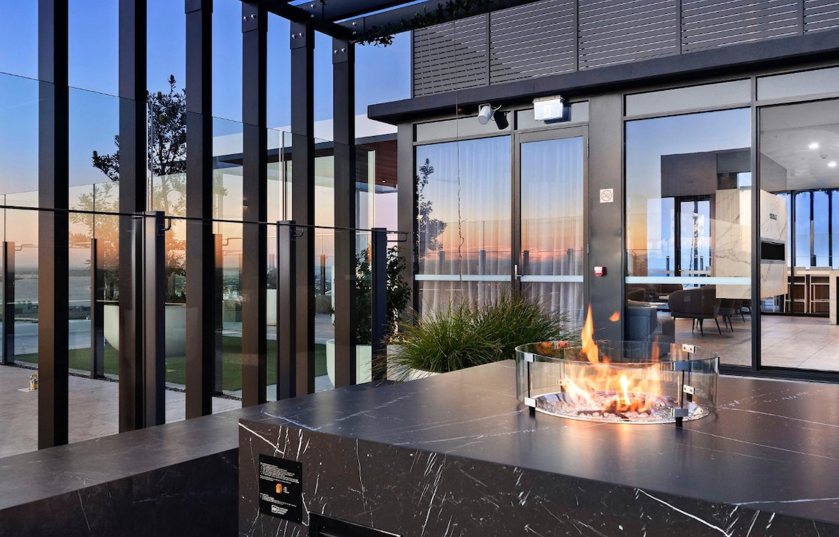 An outdoor seating area features a modern fire pit surrounded by glass barriers. The space offers an unobstructed view of the horizon, showcasing a colorful sunset. Contemporary furniture is visible in the background, blending indoor and outdoor living.