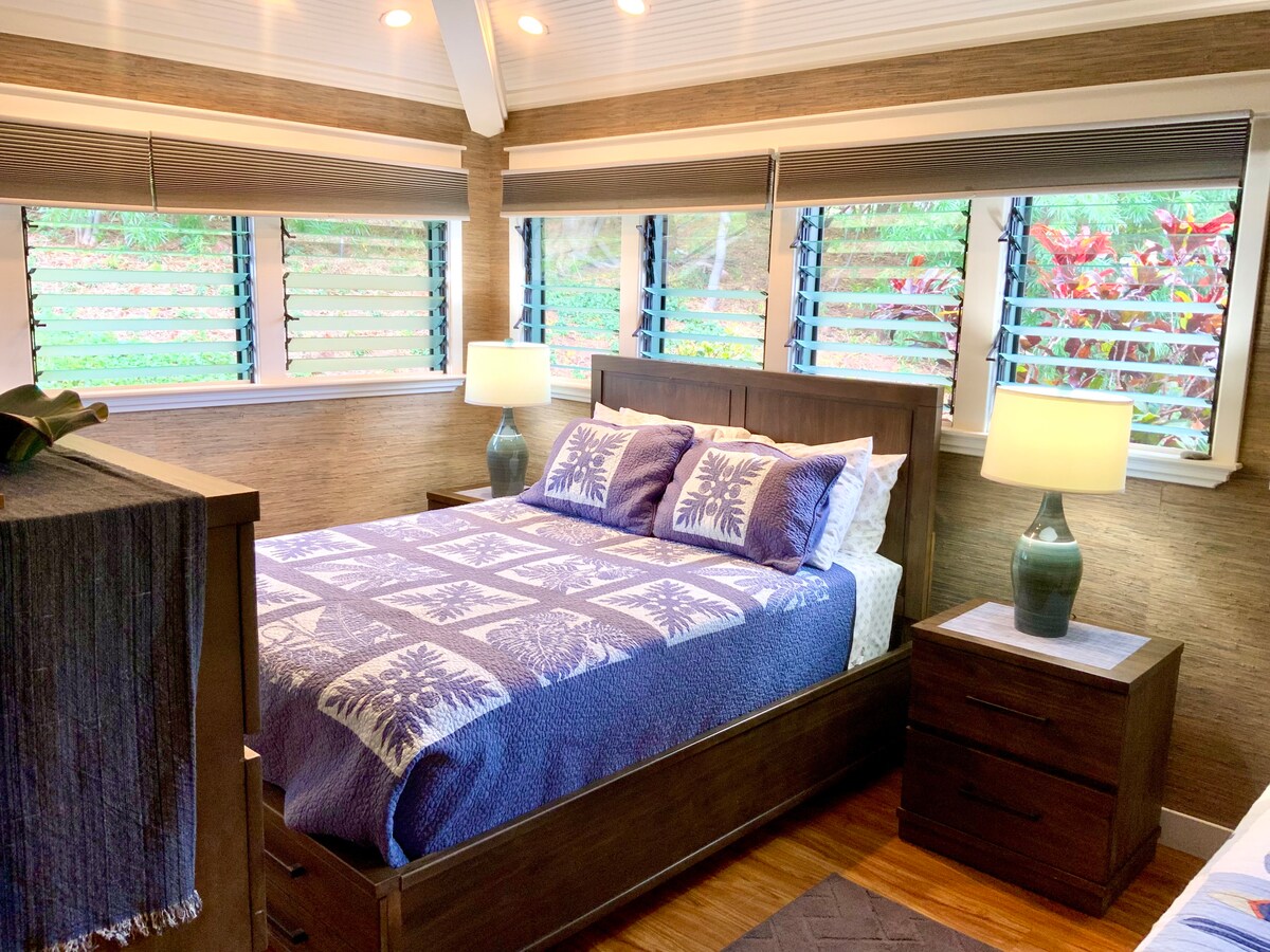 A comfortable bedroom features a spacious bed covered with a patterned blanket. Natural light filters through louvered windows, illuminating the room. Two bedside lamps provide additional lighting, and decorative plants complement the relaxing ambiance. A tasteful area rug adds texture to the hardwood floor.