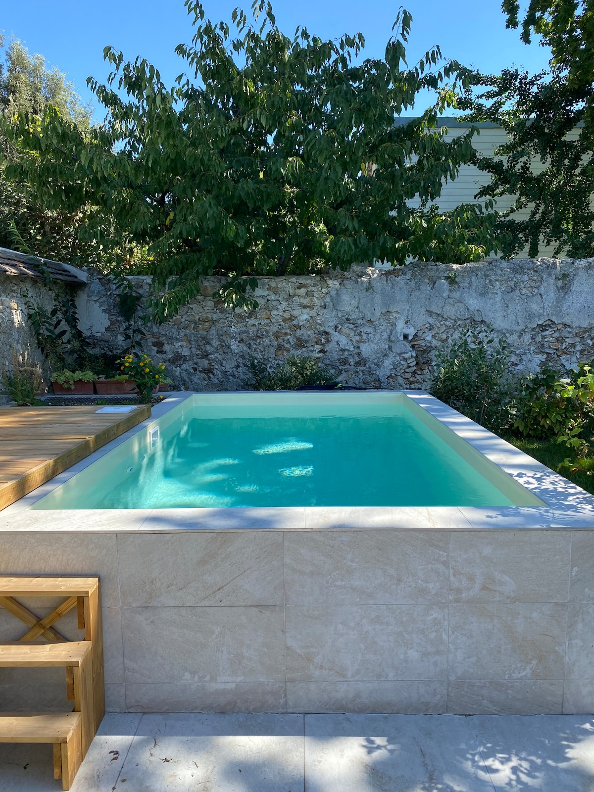 A serene pool is situated within a stone-walled garden, featuring clear turquoise water surrounded by lush greenery. Wooden decking adds a natural touch, creating a peaceful outdoor retreat.