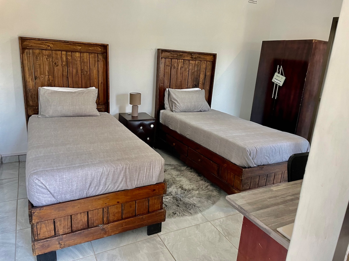 The room contains two single beds with wooden frames positioned side by side, both dressed in grey linens. A simple nightstand with a lamp sits between the beds. A dark wooden wardrobe is visible in the corner, adding functional storage space.