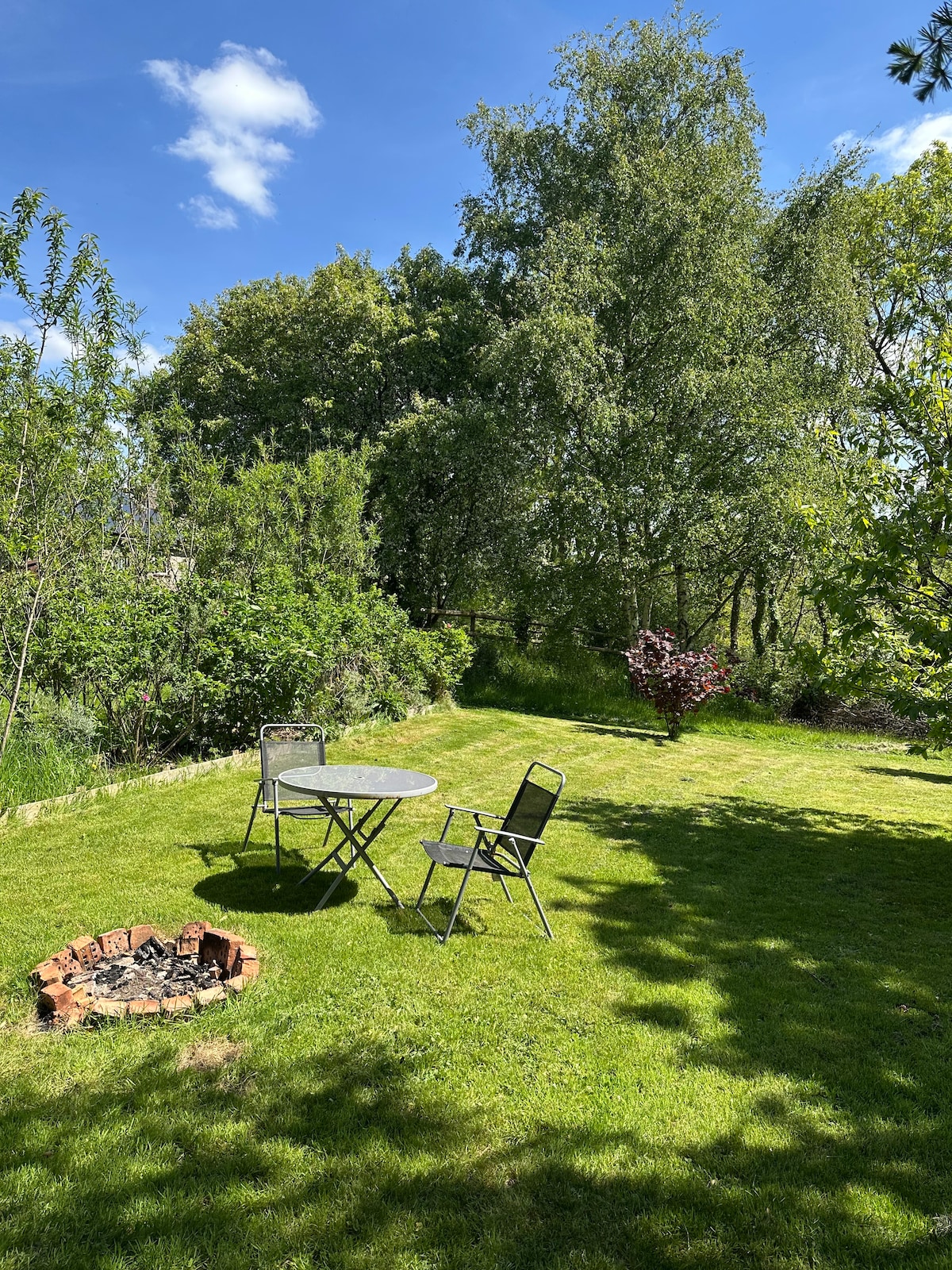 A private garden area is presented, featuring a circular stone fire pit surrounded by a grassy lawn. A compact round table with two chairs is positioned beneath the shade of trees, complemented by surrounding greenery and blue skies with scattered clouds.