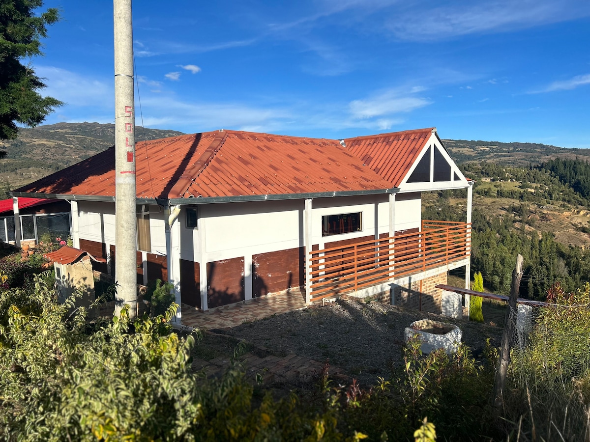 The exterior of the cabin showcases a sloping red roof and bright white walls, complemented by large windows offering expansive mountain views. A spacious terrace is visible, framed by wooden railings. Surrounding greenery blends harmoniously with the natural landscape.