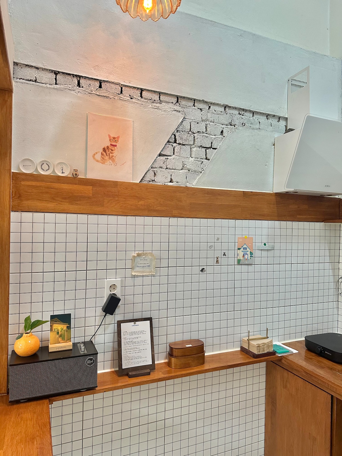 A well-organized kitchen corner features wooden shelves and a tiled backsplash. A playful cat illustration decorates the wall. Essential items include a compact printer on a dark surface and a fruit bowl with a bright orange. Note cards and a small storage box add functionality.