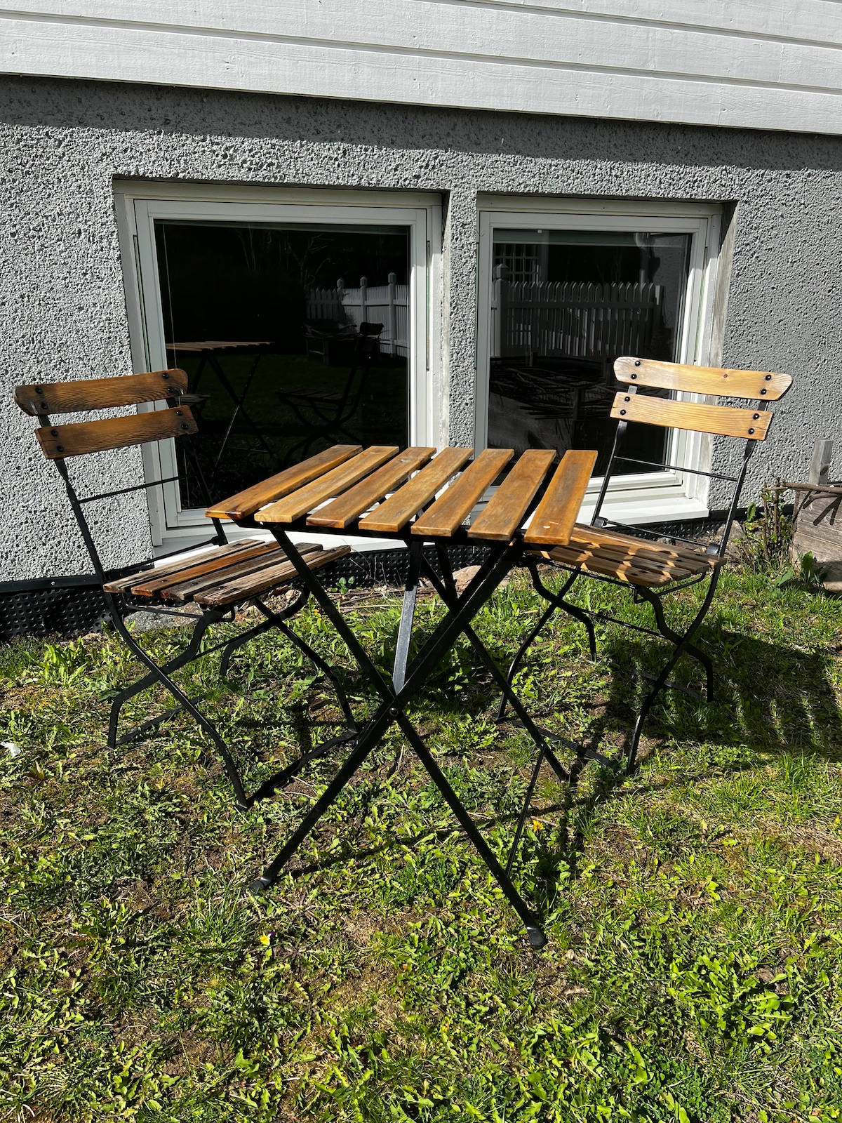 A small outdoor table with wooden slats and metal legs is positioned in a grassy area. Two matching chairs, made of wood and metal, are placed next to the table. Large windows in the background allow natural light to enter the space.