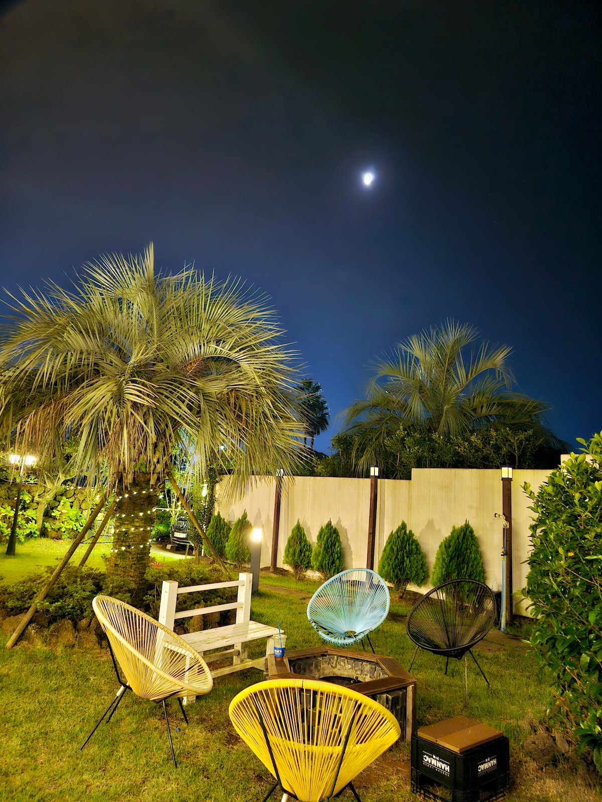 A serene outdoor space is illuminated under the night sky, featuring four comfortable chairs arranged around a small table. Lush greenery, including palm trees and hedges, provides privacy, while fairy lights delicately adorn the background, creating a relaxing ambiance for evening gatherings.