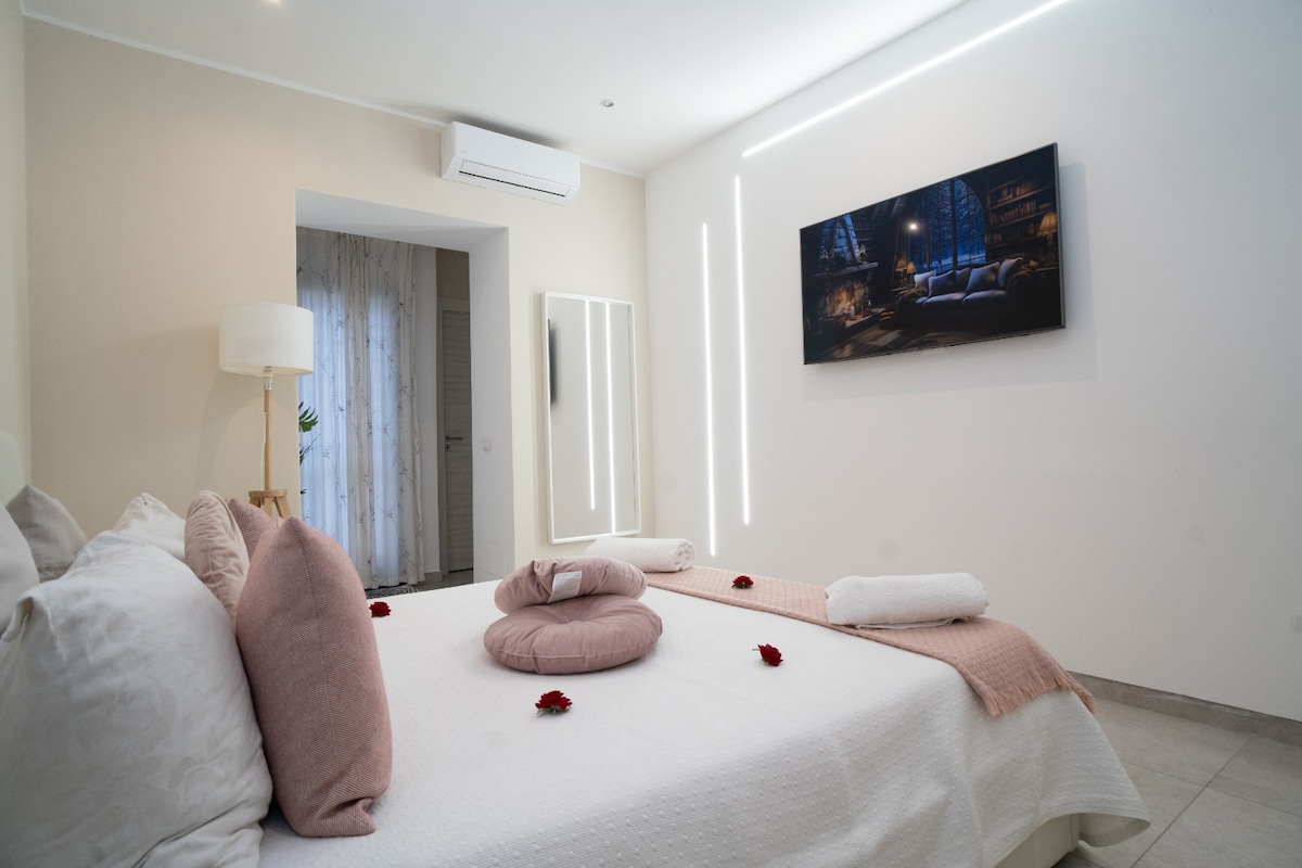 A well-lit bedroom features a spacious bed adorned with decorative pillows and towels. A large wall-mounted television is positioned above the bed, while soft lighting enhances the room's modern decor. A mirror and a light stand are visible, contributing to the room's bright ambiance.
