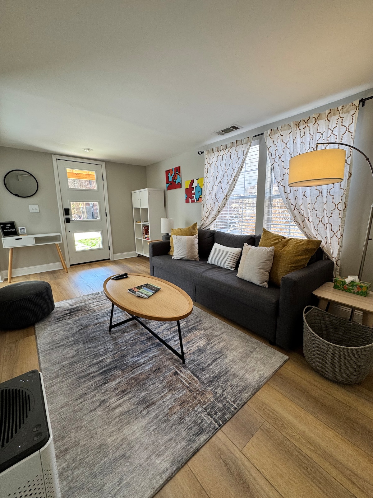 A modern living room features a gray convertible sofa adorned with colorful cushions. The space includes a round coffee table positioned on a soft area rug. Natural light filters through large windows with decorative curtains, illuminating the room's neutral tones and simple decor.