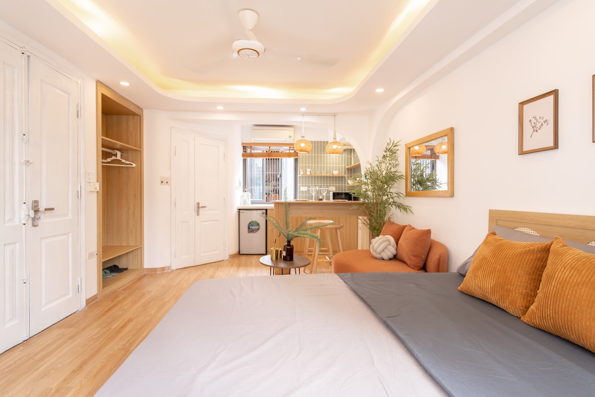 A spacious studio layout showcases a comfortable sleeping area with a standard bed featuring soft pillows. Natural light fills the room, which is complemented by a kitchenette and a cozy seating area with an orange couch. Light wood flooring adds warmth to the space.