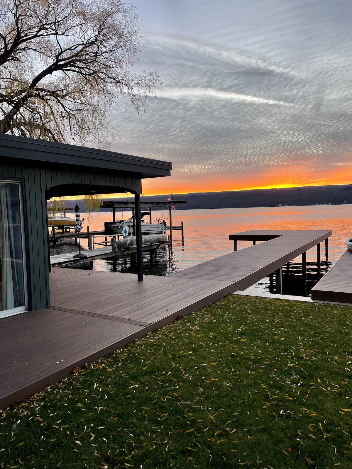 A spacious deck extends towards the calm water, framed by lush green grass. The sunset casts a warm glow over the horizon, reflecting vibrant colors on the water's surface. A dock is visible, along with silhouettes of boats at rest nearby.