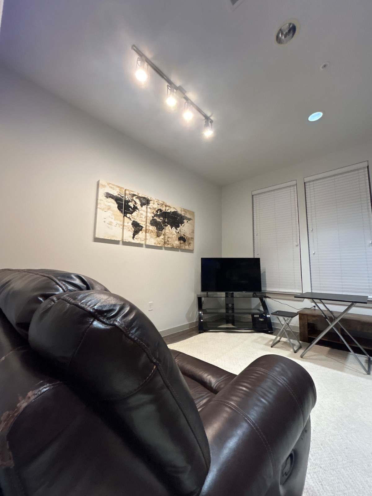 A cozy living area is featured, with a dark leather recliner positioned to one side. A wall-mounted TV sits on a glass stand opposite a table. A world map art piece adorns the wall, while natural light filters through partially closed white blinds.