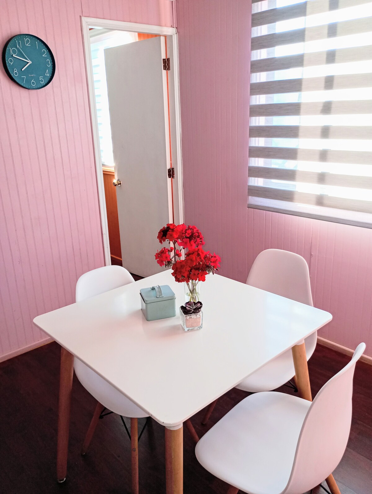 A dining area is featured, showcasing a white table surrounded by four modern chairs. A small vase with red flowers is placed at the center of the table. Light streams in through partially closed shades, highlighting the pink walls and a clock on the wall.