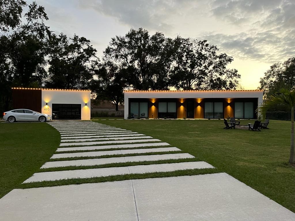 The modern guesthouse is set against a spacious green lawn, with a paved walkway leading to the entrance. The structure features multiple glass windows, illuminated softly by exterior lights. A seating area with chairs is visible on the lawn, creating an inviting outdoor space.