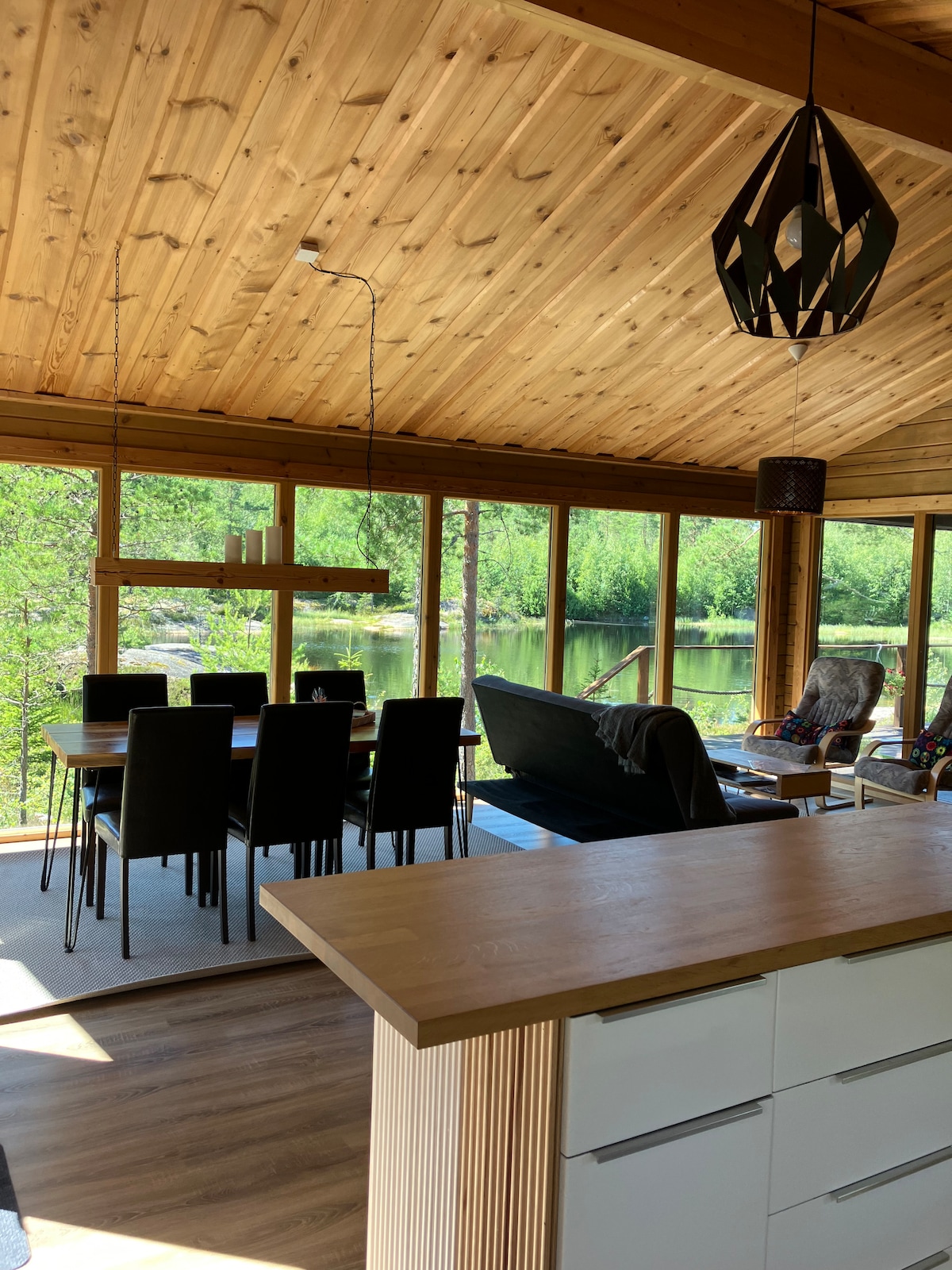 An open living area features high wooden ceilings and large windows that provide views of the surrounding nature and lake. A dining table with dark chairs sits beside the kitchen, while a comfortable seating area is arranged for relaxation.