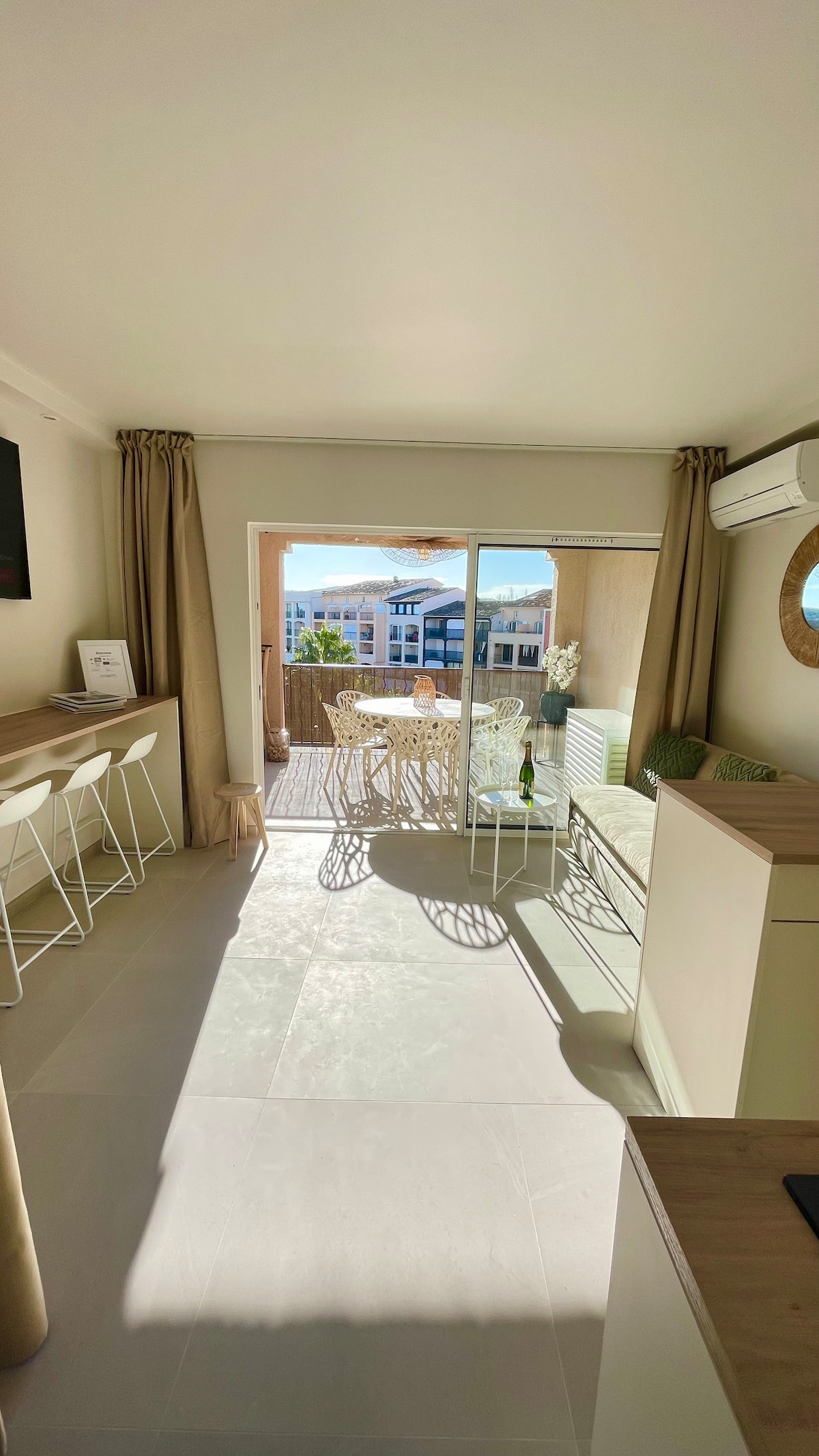 A bright living area features an open layout with large sliding glass doors that lead to a terrace. Light-colored walls complement modern furnishings, including a compact kitchen and bar stools. A cozy dining set awaits on the terrace, showcasing views of the surrounding area.