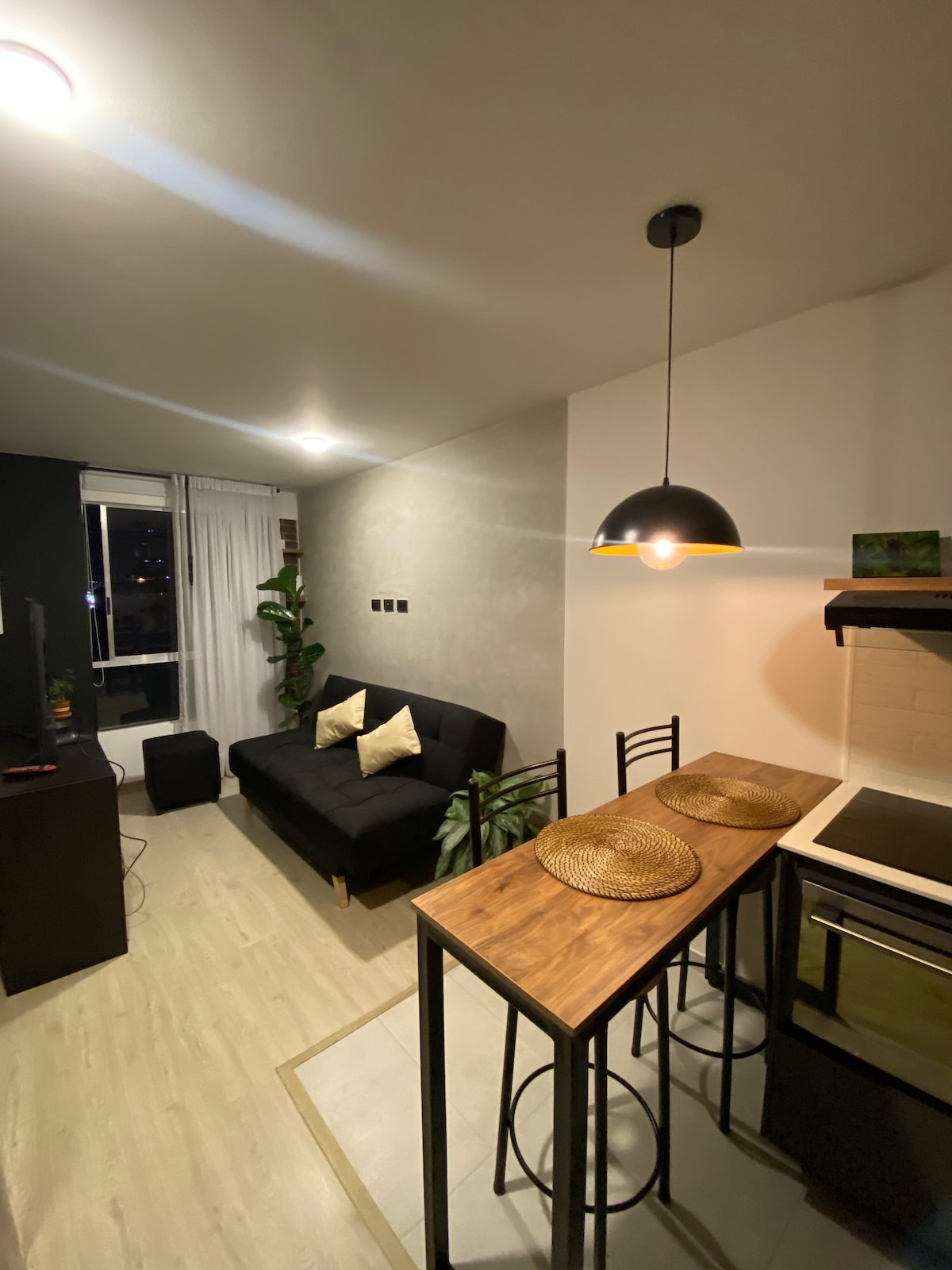A modern living space showcases a dark sofa with decorative cushions and a tall, seated breakfast area adjacent. A wooden table is set with circular placemats, while the kitchen features sleek cabinetry and a stove. Soft lighting adds a comfortable ambiance to the room.