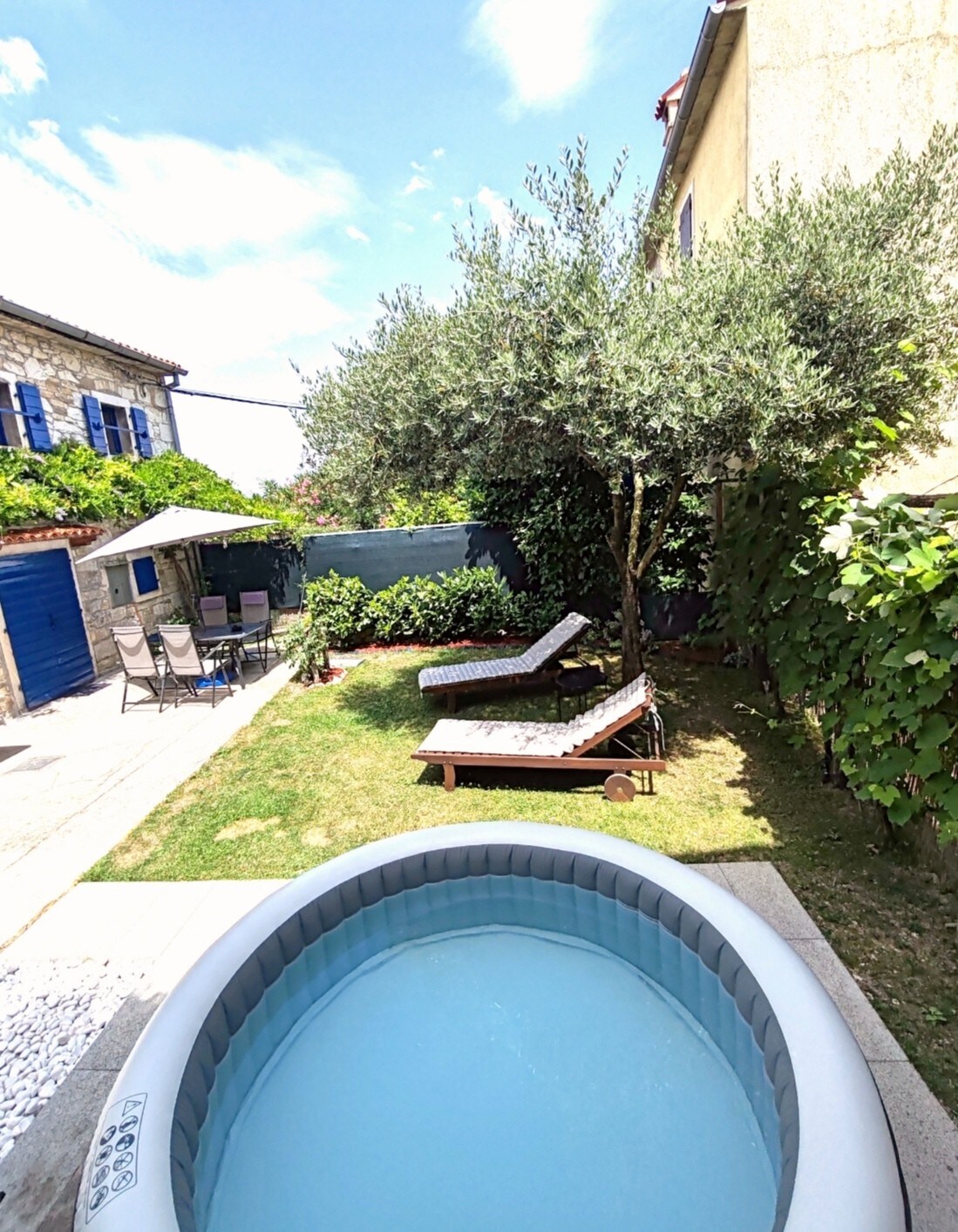 A private, inflatable hot tub is situated on a stone patio, surrounded by lush greenery. Nearby, two sun loungers are positioned on a grassy area under a small tree, with a shaded outdoor dining table and chairs visible in the background.