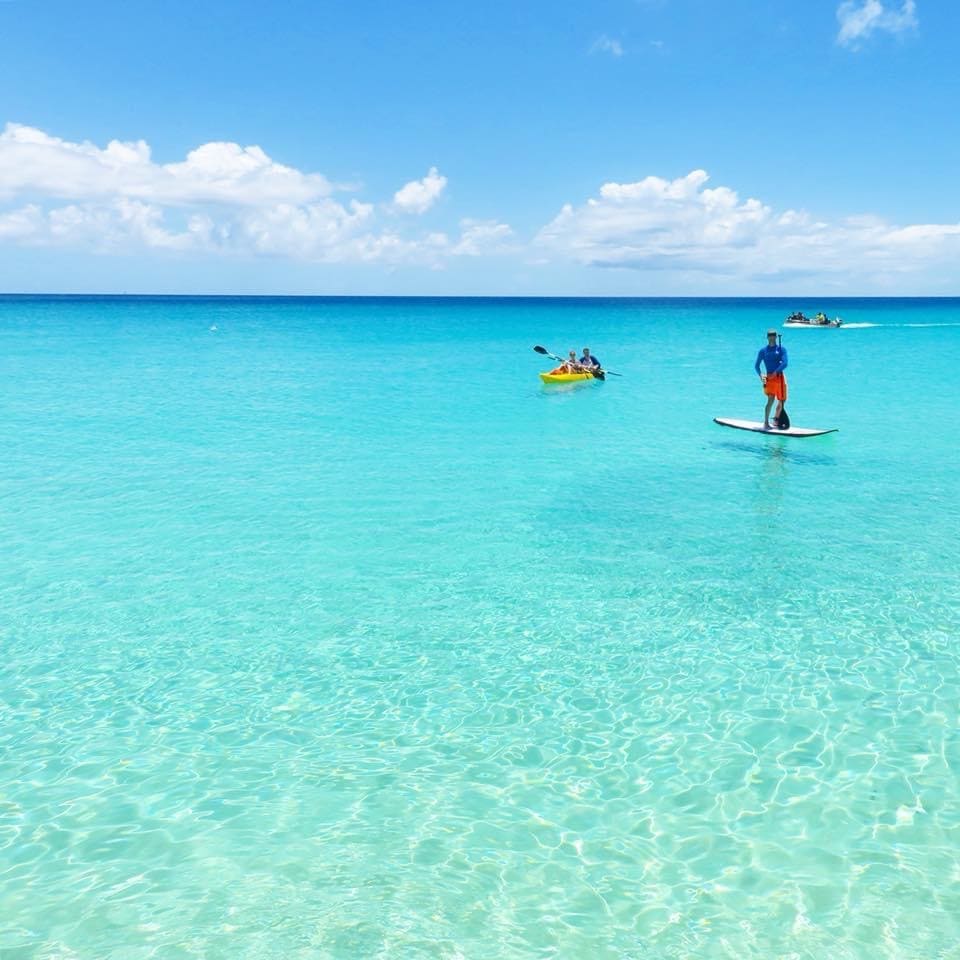 Clear turquoise waters stretch into the horizon, where bright blue skies and fluffy white clouds can be seen. A person stands on a paddleboard, while a kayak with two people can be spotted nearby, creating a scene of serene outdoor activity.