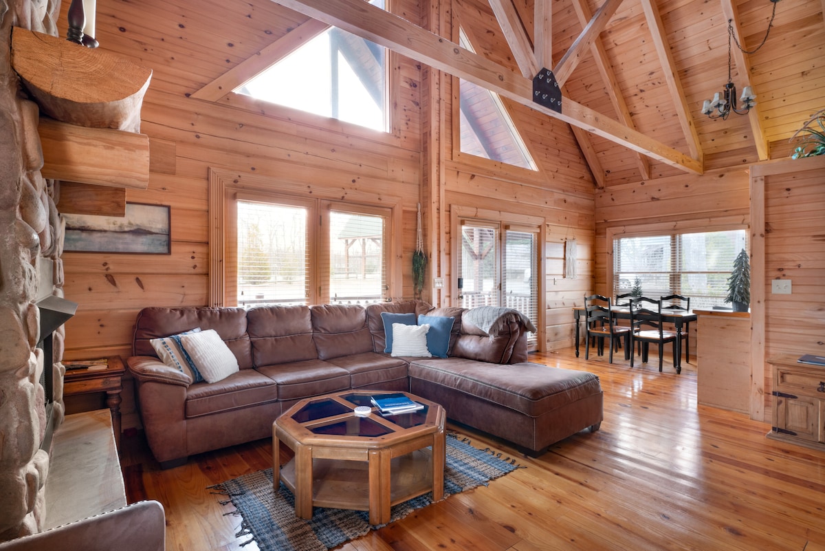 A spacious living room features a large sectional sofa arranged around a wooden coffee table. Natural light floods the area through tall windows, highlighting the vaulted wood ceiling. A cozy fireplace is visible, along with a dining area in the background.