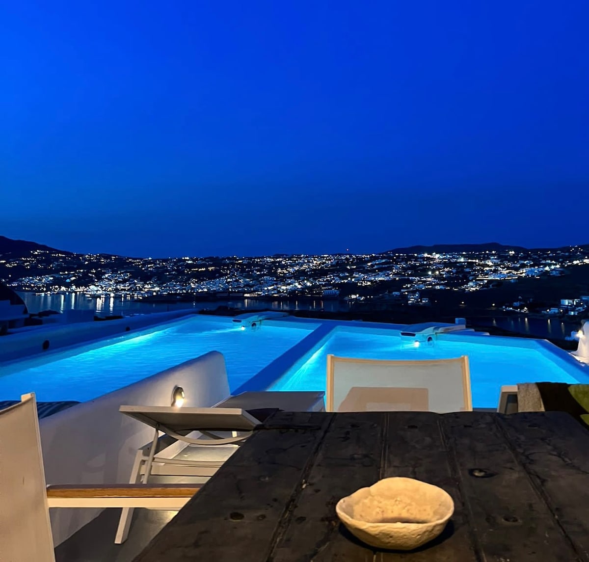 An outdoor terrace features a wooden table with a bowl positioned on top. In the background, two illuminated pools reflect the evening sky, showcasing views of the distant city lights that twinkle against a deep blue backdrop.