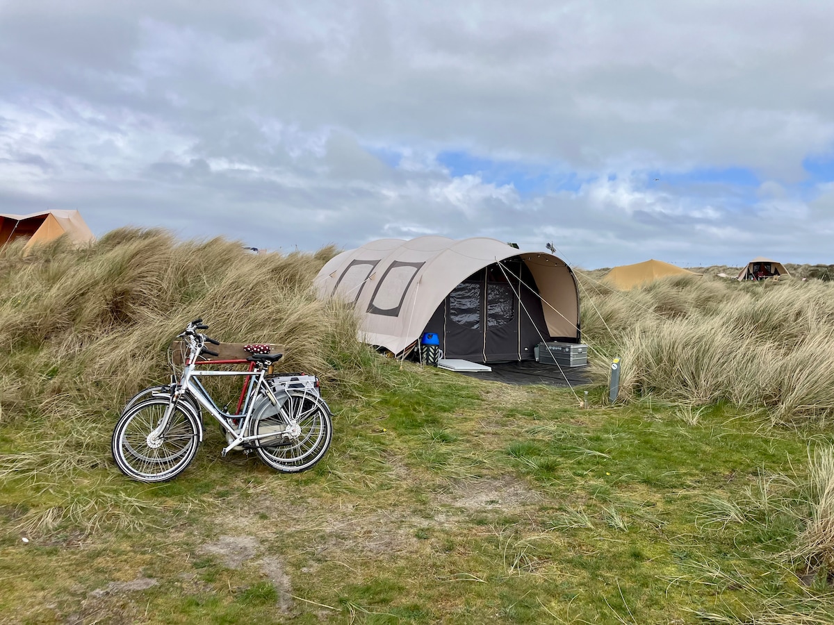 A spacious tent is positioned among natural dunes, characteristic of the campsite's landscape. Two bicycles are parked nearby on a grassy path leading to the tent entrance, which is partially opened, inviting exploration into the interior.