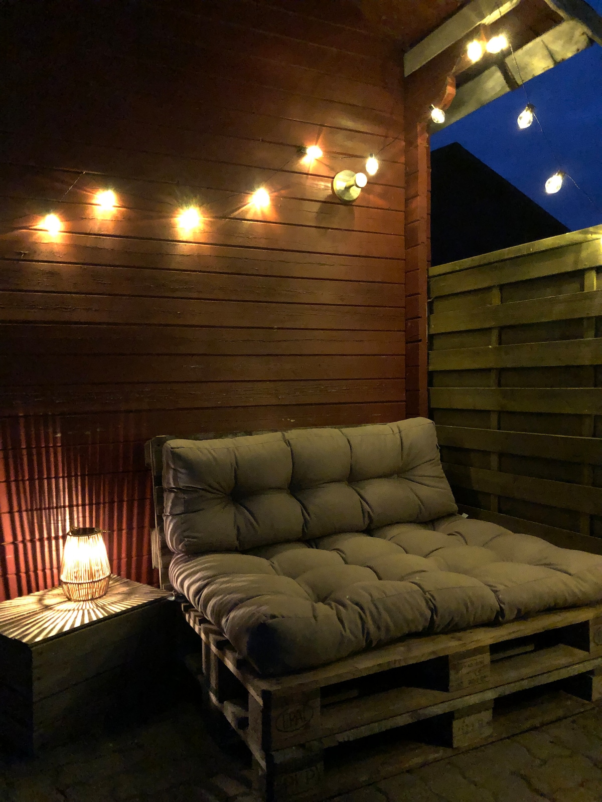 A cozy outdoor seating area is displayed with a cushioned sofa made from wooden pallets, illuminated softly by string lights. A lantern on a small wooden table provides additional warm light against the wooden wall that surrounds the space.