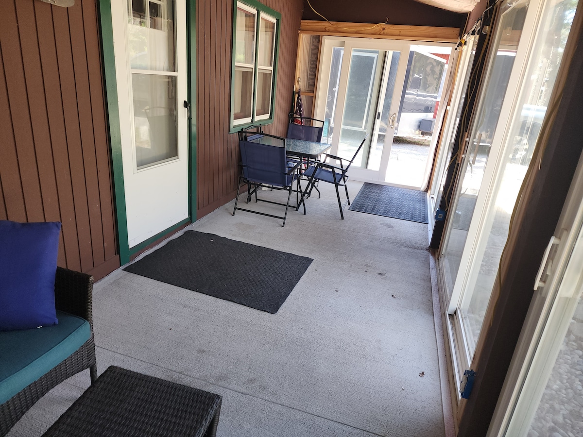 A covered porch area features a table and chairs, providing an inviting space for relaxation. The flooring is gray and smooth, leading to glass doors that open to the outdoors. Two seating areas are visible, complemented by window views and a neutral color scheme.