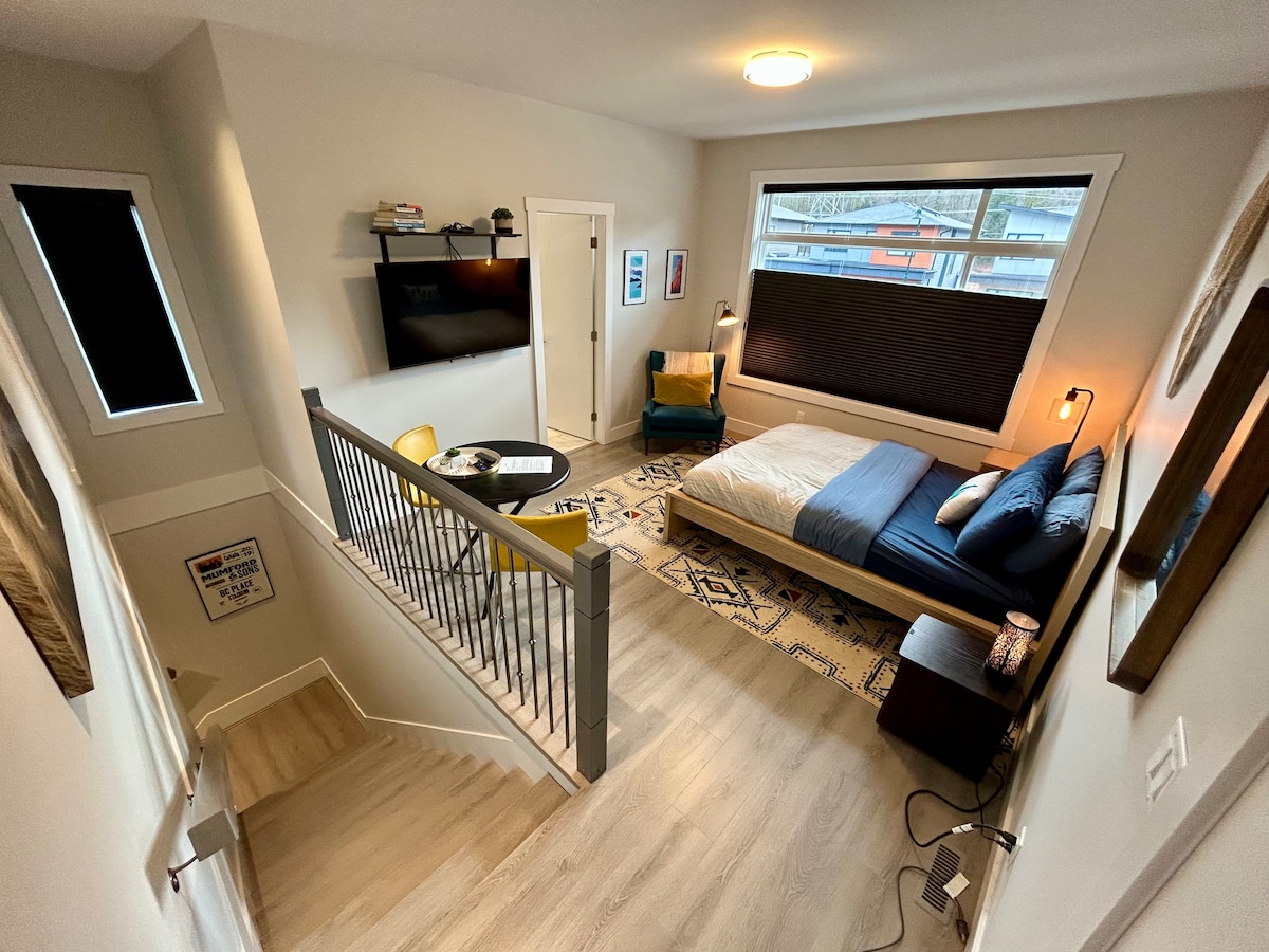 An airy studio space is captured from above, showcasing a cozy bed with blue and white linens. A comfortable chair sits adjacent to the bed, and a television hangs on the wall. Large windows allow natural light to illuminate the room.