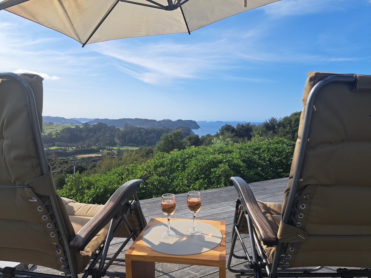 Two comfortable chairs are positioned on a deck, facing a scenic view of the coastline and rolling hills. A small table in between holds two glasses of rosé, with an umbrella providing shade against the bright blue sky.