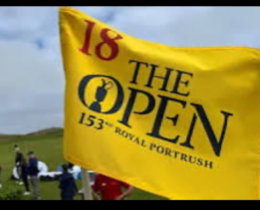 A bright yellow flag is prominently displayed, featuring the logo and text for 'The Open,' indicating the 18th hole at Royal Portrush. Soft green grass is visible in the background, along with indistinct figures engaging in activities nearby.