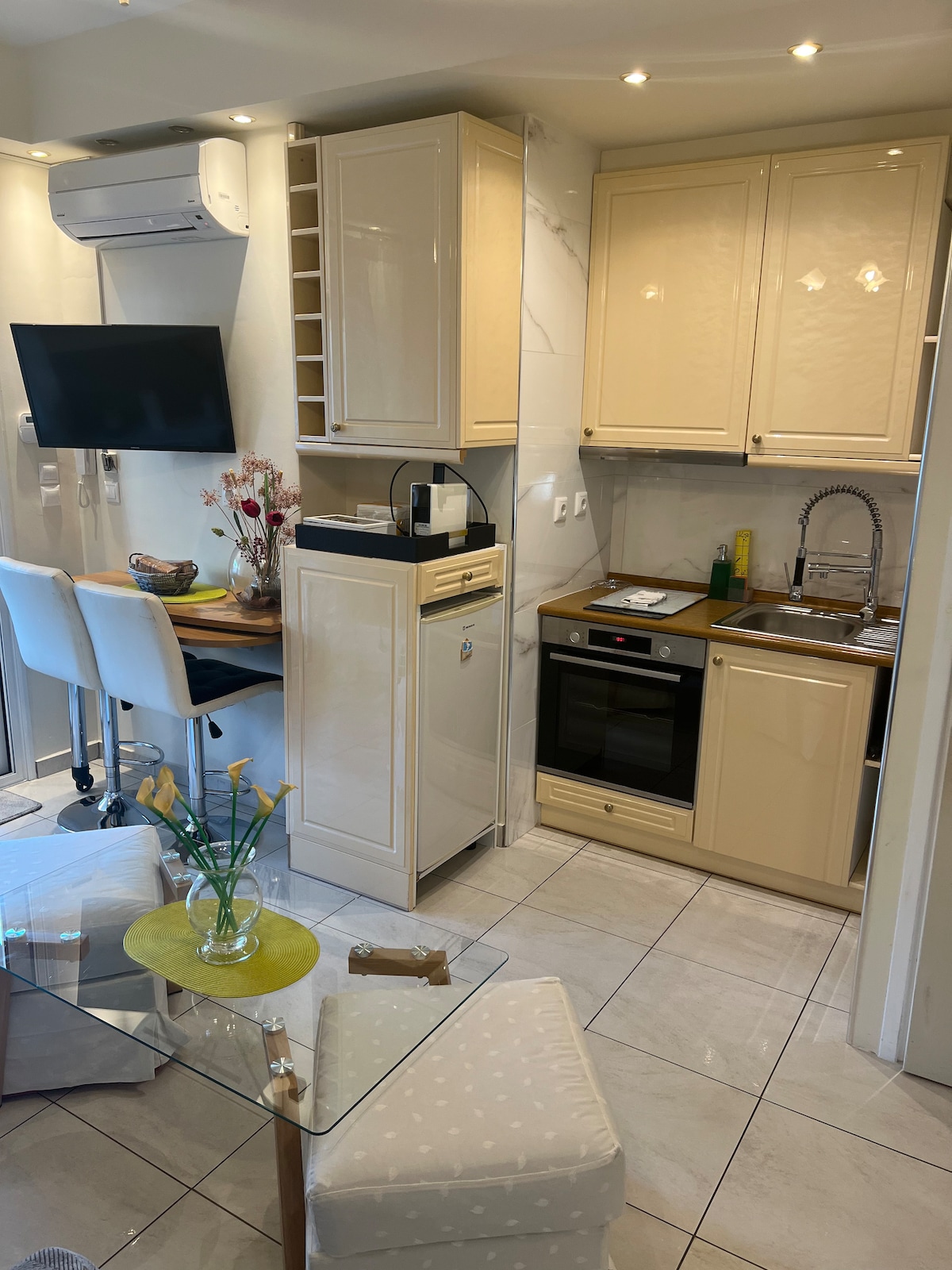 The kitchenette features modern appliances, including an oven and refrigerator. A small dining area with two stools is present, along with decorative elements such as flowers on a round table. Light fixtures enhance the space, complementing the beige cabinetry and tiled floor.