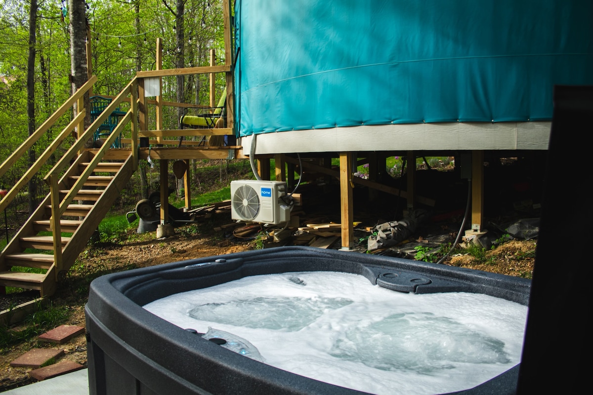 A hot tub is positioned in the foreground, showcasing bubbling water. In the background, a wooden staircase leads up to the yurt, surrounded by lush greenery. An air-conditioning unit is mounted beneath the elevated structure.