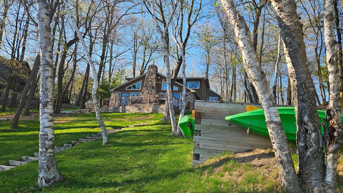 The image shows a lakefront home surrounded by tall trees, with steps leading up to the entrance. The house features multiple large windows and stone accents on the exterior. Two bright green kayaks are positioned on a wooden platform, ready for use.