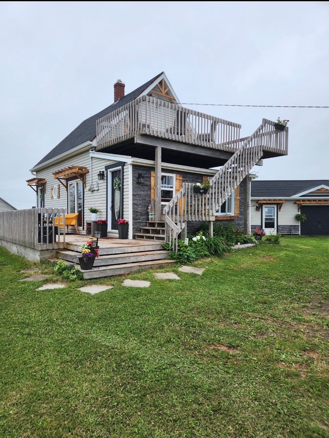 A charming two-story house features a welcoming deck with wooden railings and flower planters. A spiral staircase leads to the upper deck, surrounded by a grassy area and stone path. Soft colors of the exterior complement the natural surroundings.
